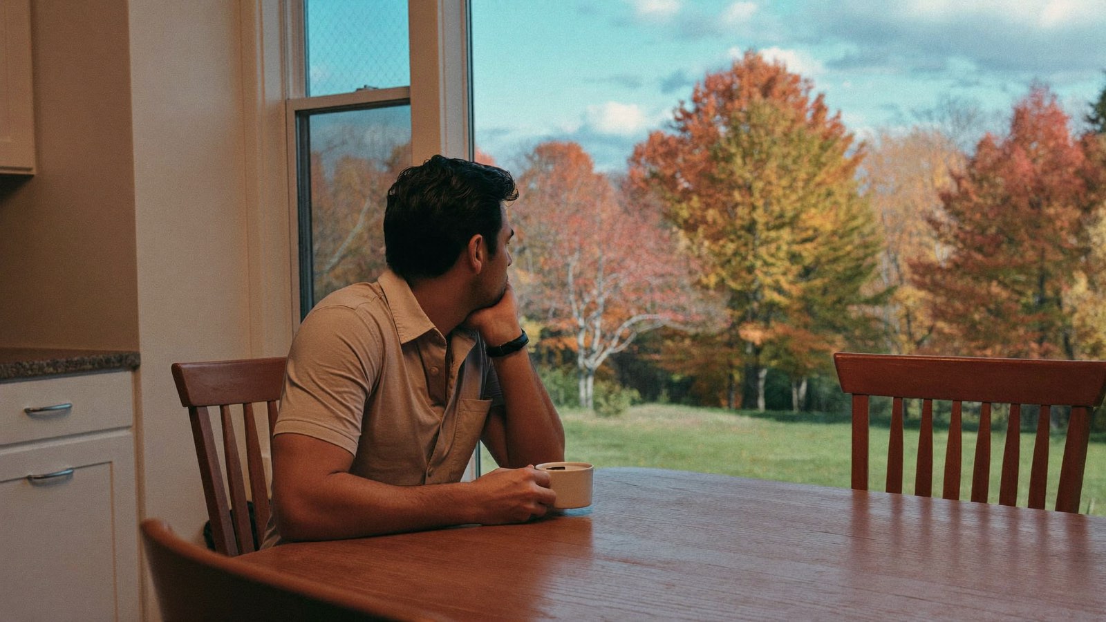 Man in his late 30s with thick dark hair sitting at a wooden kitchen table with a coffee cup, looking out a window at autumn trees