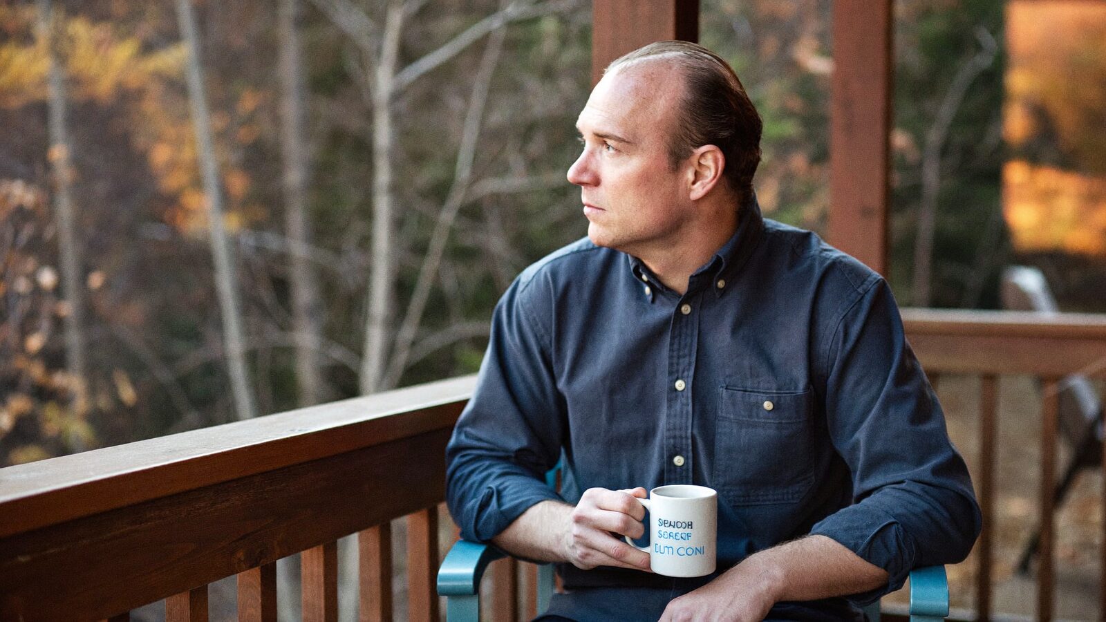 Man in his late 30s with a slightly receding hairline sitting on a wooden porch with a coffee mug, autumn trees behind him