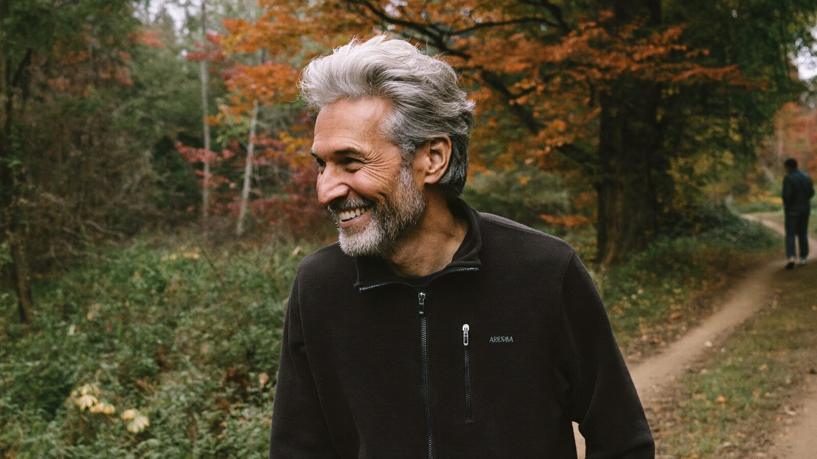 Man in his late 40s with a full, natural hairline walking along a lakeshore path in the evening sun, smiling